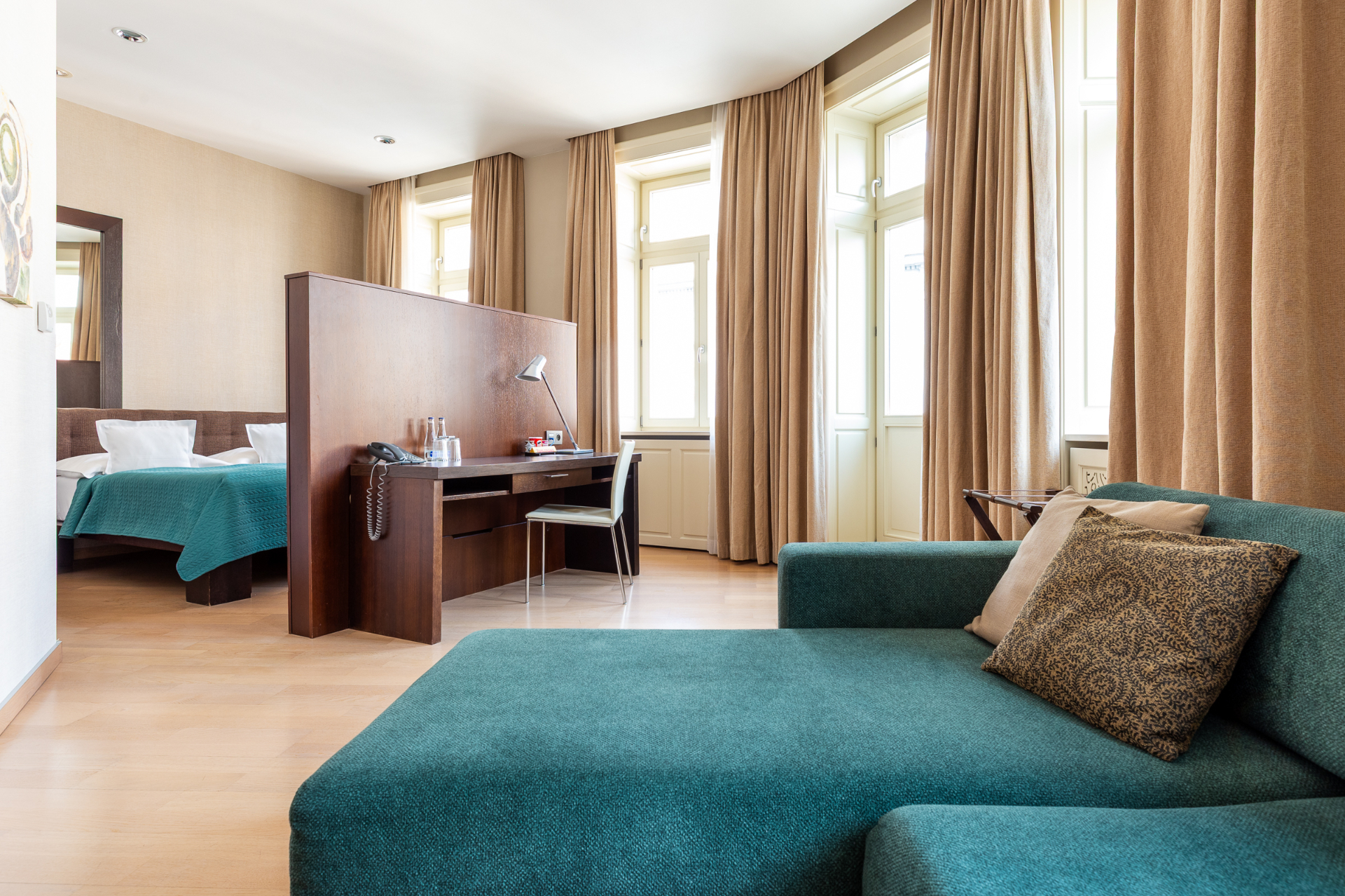 Deluxe Suite with teal chaise lounge, desk divider, bedroom and tall arched windows